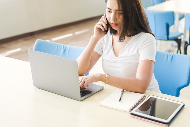 Young asian woman talking with someone on mobile phone while working with laptop.