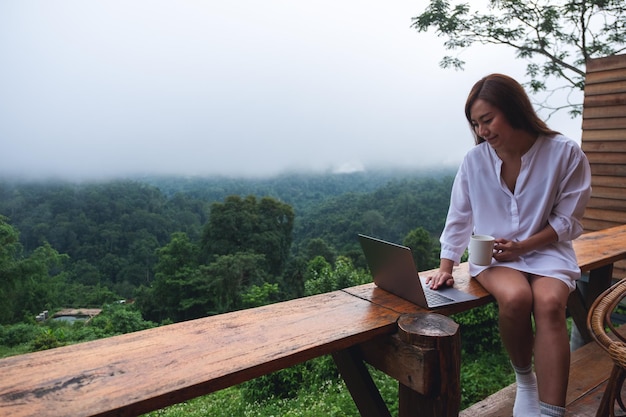 A young asian woman using and working on laptop computer while sitting on balcony with a beautiful nature view on foggy day