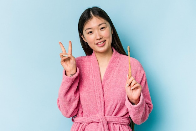 Young asian woman wearing a bathrobe brushing teeth isolated on pink background joyful and carefree showing a peace symbol with fingers