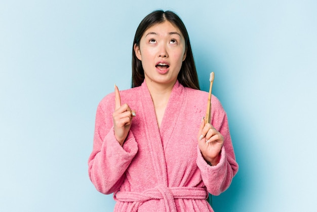 Young asian woman wearing a bathrobe brushing teeth isolated on pink background pointing upside with opened mouth