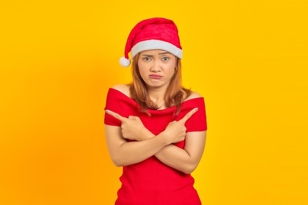 Young Asian woman wearing Christmas hat pointing to copy space with finger and having doubts