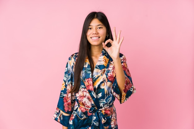 Young asian woman wearing a kimono pajamas cheerful and confident showing ok gesture.