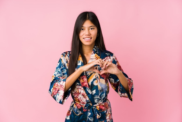 Young asian woman wearing a kimono pajamas smiling and showing a heart shape with hands.