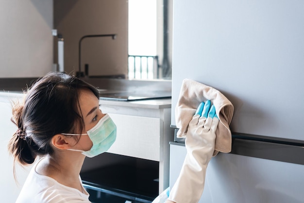 Young Asian woman wearing medical mask and hands in rubber white glove cleaning refrigerator with napkin in kitchen at home Closeup