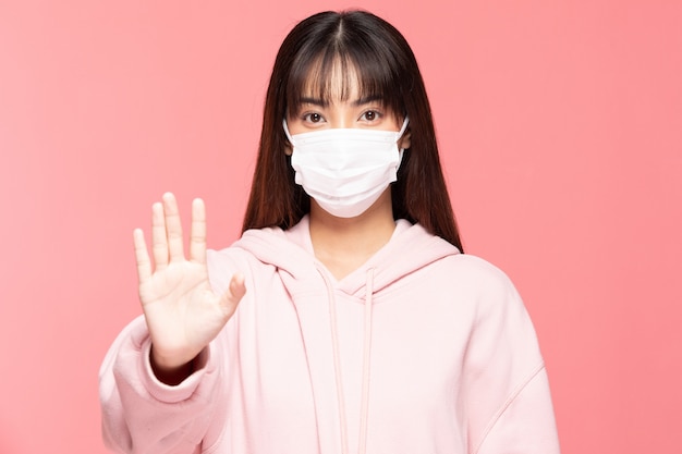 Young asian woman wearing protection mask or surgical mask for protected virus and air pollution making hand stop virus on pink wall,Health care and coronavirus concept