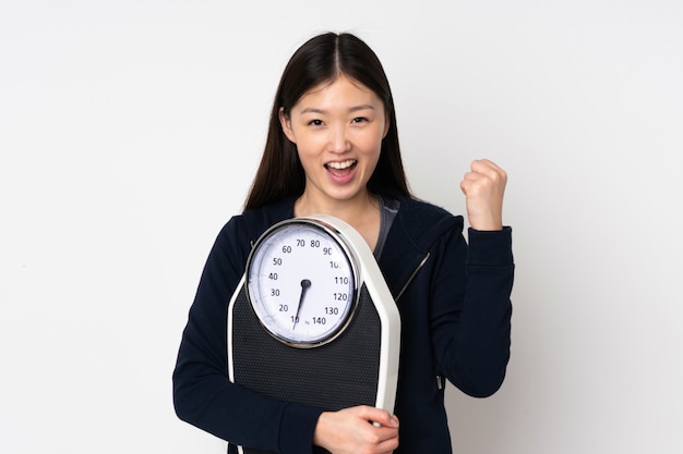 Young asian woman on white wall with weighing machine and doing victory gesture