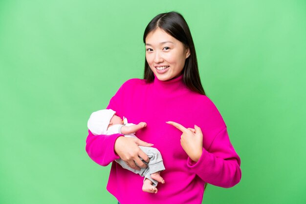 Young Asian woman with her newborn baby over isolated chroma key background and pointing it