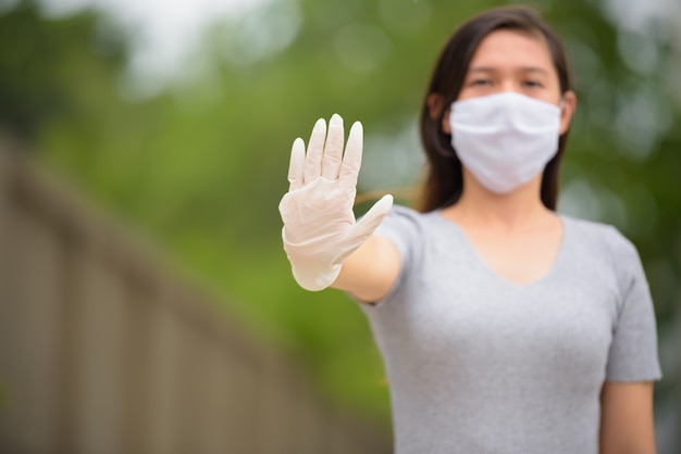 Young Asian woman with stop gesture while wearing mask and gloves outdoors