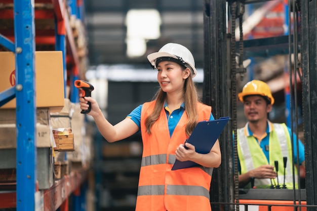 Young Asian woman worker using barcode scanner checking goods and box shelf stock in the warehouse factory store and smile happy coworker man drive forklift in back Warehouse Logistic concept