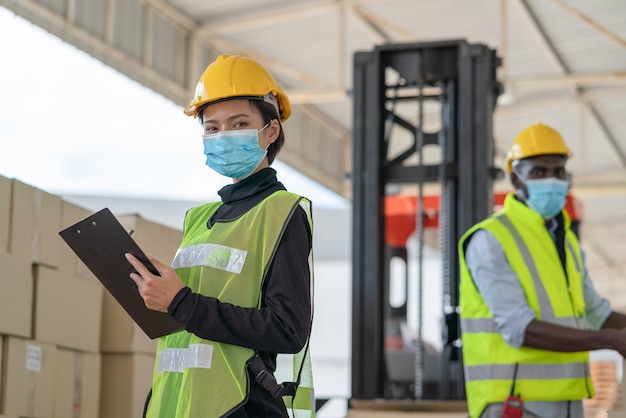 Young Asian woman worker with man workers wear face mask for protect coronavirus working at logistics warehouse factory