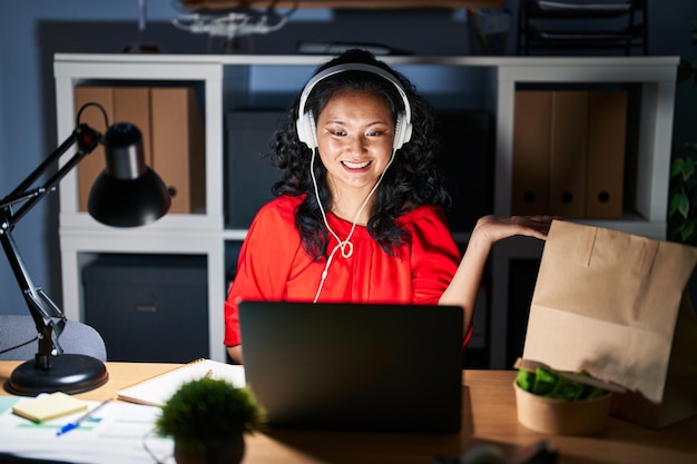 Young asian woman working at the office with laptop at night smiling cheerful presenting and pointing with palm of hand looking at the camera