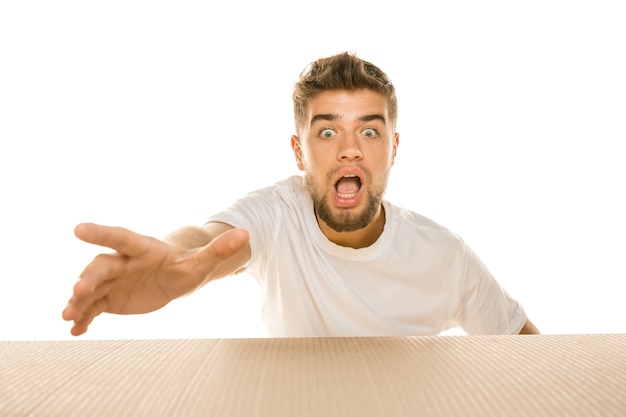 Young astonished man opening the biggest postal package isolated on white. Shocked male model on top of cardboard box looking inside. 