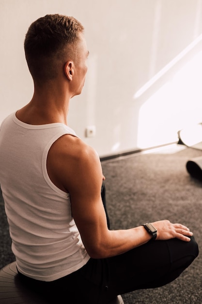 A young athlete in a white tshirt sits near the window and looks into the distance