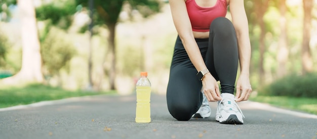 Young athlete woman tying running shoes with Energy Drink water female runner ready for jogging outside asian Fitness walking and exercise in the park morning wellness wellbeing and sport concepts