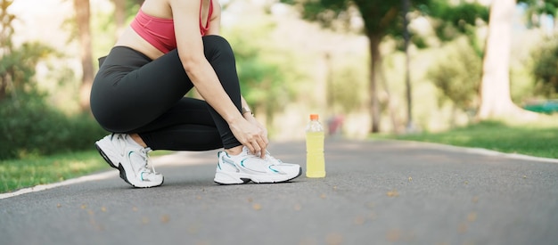 Young athlete woman tying running shoes with Energy Drink water female runner ready for jogging outside asian Fitness walking and exercise in the park morning wellness wellbeing and sport concepts