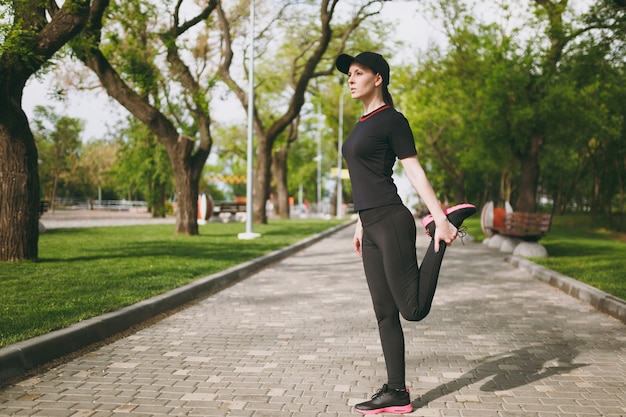 Young athletic beautiful brunette woman in black uniform, cap doing sport stretching exercises, warm-up before running or training, standing in city park outdoors