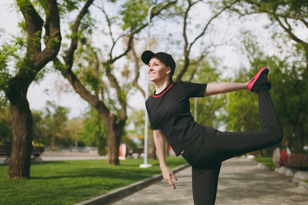 Young athletic beautiful brunette woman in black uniform, cap doing sport stretching exercises, warm-up before running or training, standing in city park outdoors