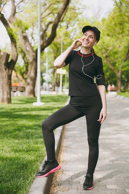 Young athletic beautiful brunette woman in black uniform and cap with earphones listening to music, standing before running, training on path in city park outdoors