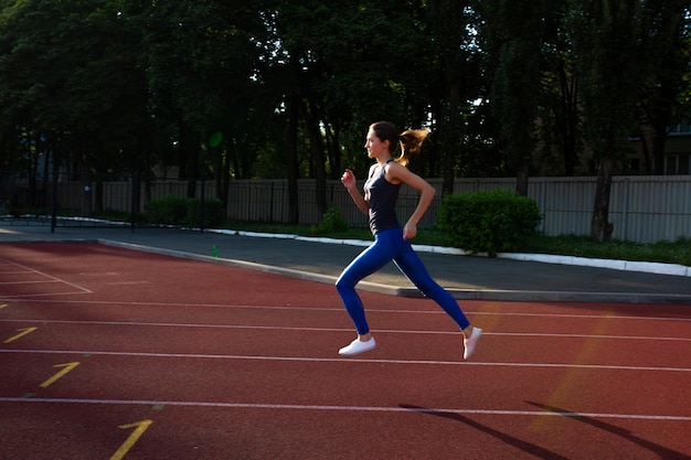 Young athletic girl in grey t shirt and blue leggings running during sunny evening on stadium track. Space for text