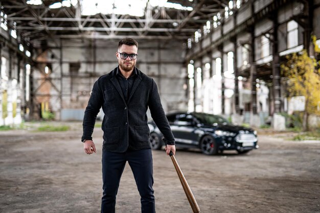 Young athletic guy in jeans holding a baseball bat Brutal image Bearded man in sunglasses Fashion car on the background