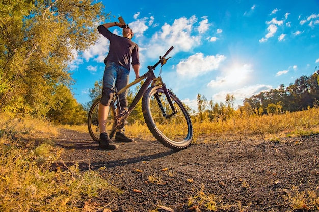 Young athletic man in a black T-shirt, blue jeans shorts and knee pads on a sports bike drinks water from a shaker on an colorful autumn with a bright sky.