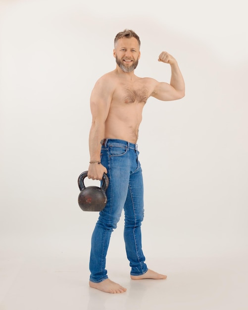 Young athletic man with naked torso standing holding kettlebell and straining muscles white backgrou