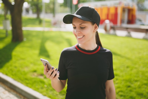 Young athletic smiling beautiful brunette girl in black uniform and cap using mobile phone during training, looking on smartphone, standing in city park outdoors