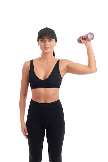 Young athletic woman in black sports wear and cap standing on white and holding dumbbell