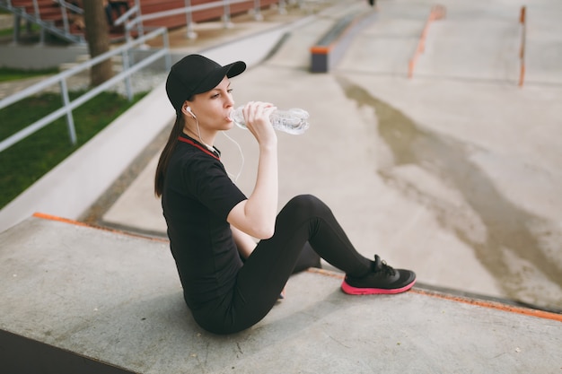 Young athletic woman in black uniform, cap with headphones listening to music, holding bottle, drinking water sitting before or after running, training in city park outdoors