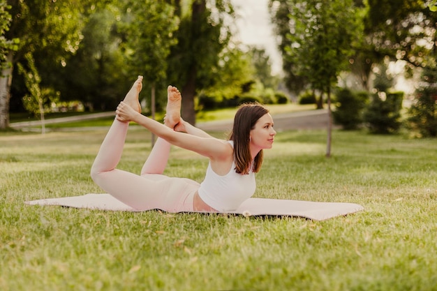 Young athletic woman practice yoga on gymnastic mat in green park on grass Stand in bow pose in sportswear and stretch muscles back pain yoga Concept of healthy lifestyle and harmony with body