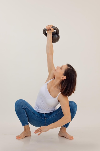 Young athletic woman sitting and lifting up kettlebell white background keeping fit by strength work