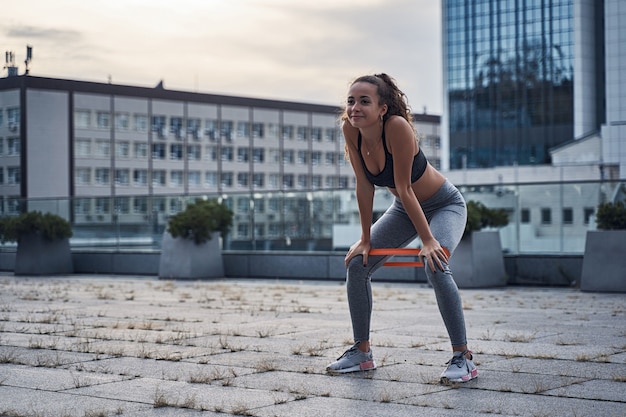 Young athletic woman smiling while doing squats exercises with sports rubber at the urban city