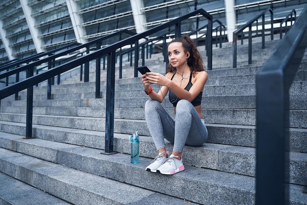 Young athletic woman in sport clothes sitting on stairs of urban stadium with phone in hands posting her training results