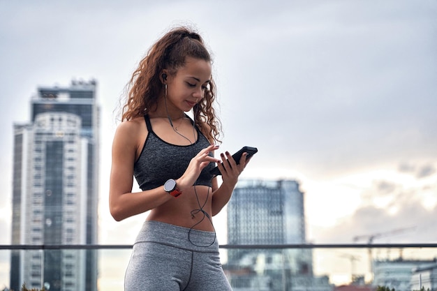 Young athletic woman in sport clothes standing with phone in hands posting her training results and listening music