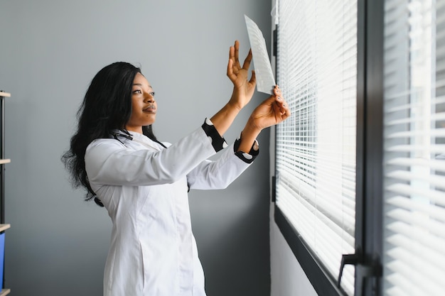 Young attractive african female doctor radiologist looking at xray image while standing in light modern office in clinic