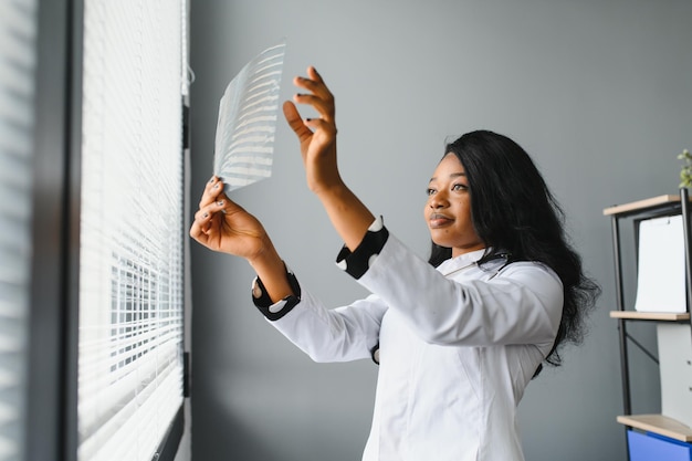 Young attractive african female doctor radiologist looking at xray image while standing in light modern office in clinic