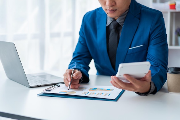 Young Attractive Asian businessman using calculator when working with computer laptop with financial chart