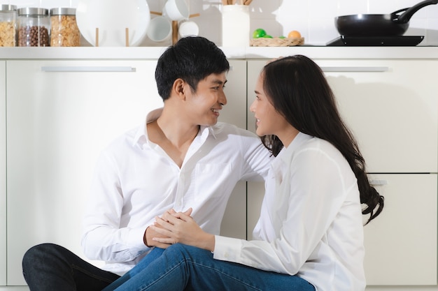 Young attractive Asian couple wearing white shirt and jeans sitting together in white kitchen