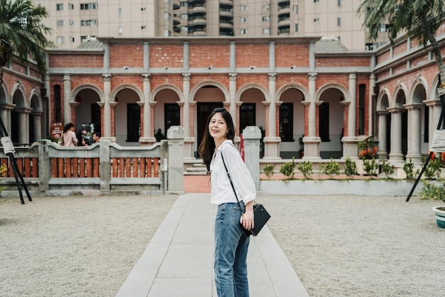 young attractive asian korean girl standing in front ground of traditional taiwanese old house. beautiful lady tourist face camera smiling and going to visit sanheyuan in Taiwan. sun flare day.