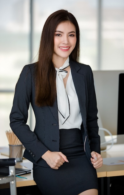 Young attractive Asian woman in black business suit sitting on table with confidence in modern looking office with blurry windows background. Concept for modern office lifestyle.