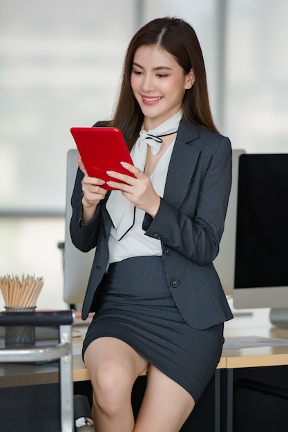 Young attractive Asian woman in black business suit working on red tablet in modern looking office with blurry windows background. Concept for modern office lifestyle.