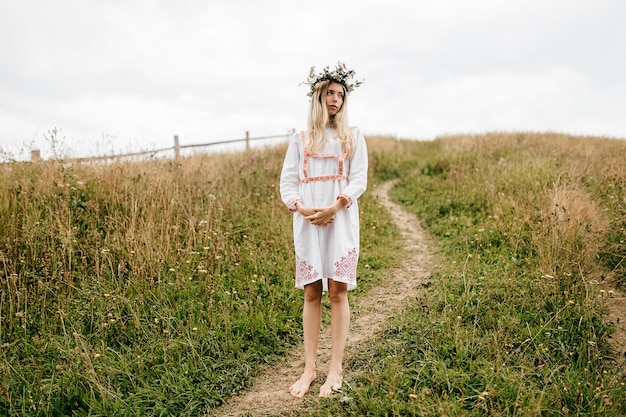 Young attractive barefoot blonde girl in white dress with ornament and flower wreath on the head posing in the field