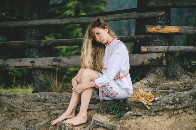 Young attractive barefoot blonde girl in white dress with ornament sitting near wooden fence outdoors