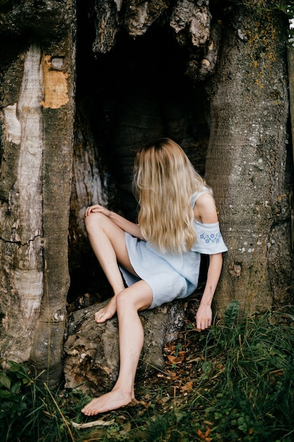 Young attractive barefoot blonde woman blue romantic dress sitting in the old tree