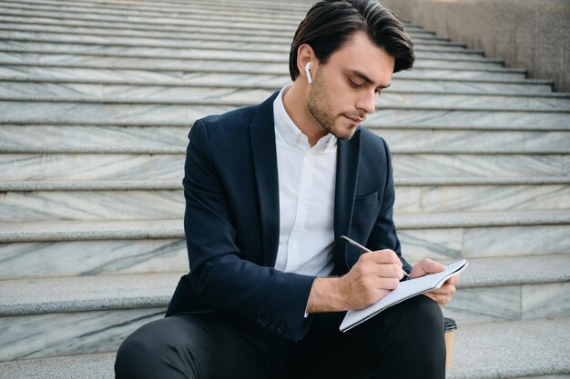 Young attractive bearded brunette man in white shirt and classic suit with wireless earphones sitting on stairs outdoor thoughtfully writing notes in notepad alone