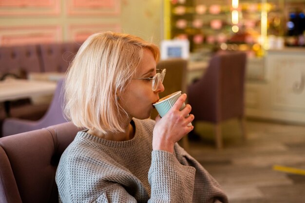 Young attractive blonde girl in cafe drinks coffee from a paper cup and relaxes