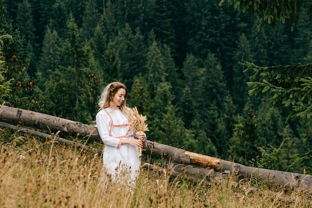 Young attractive blonde girl in white dress with embroidery posing with spikelets bouquet in the meadow over picturesque landscape