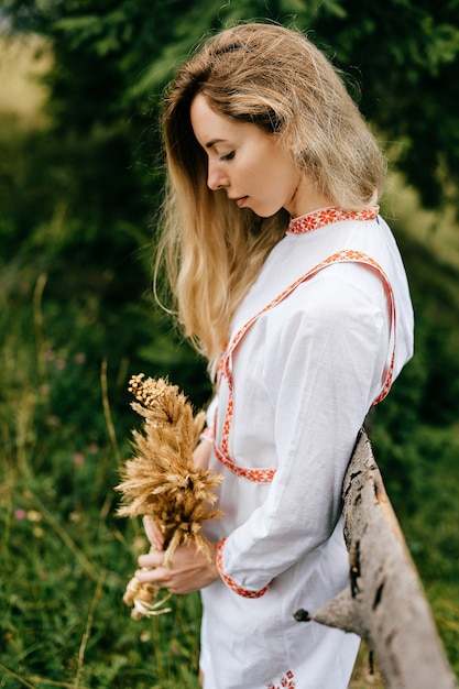 Young attractive blonde girl in white dress with embroidery posing with spikelets bouquet near wooden fence