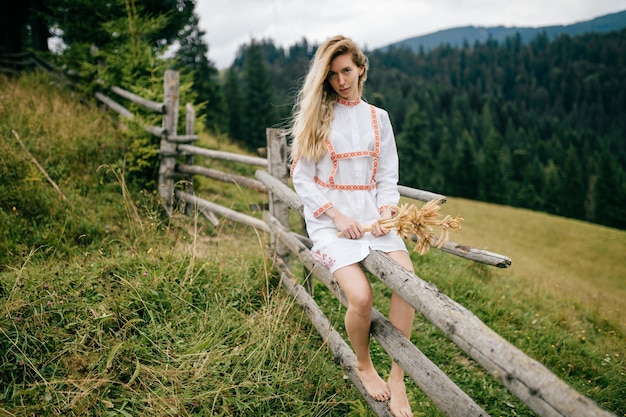 Young attractive blonde girl in white dress with ornament sitting on wooden fence with spikelets bouquet over picturesque countryside landscape