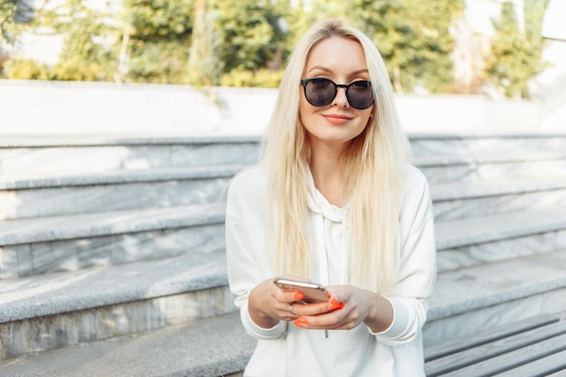 Young attractive blonde woman in sunglasses using smartphone while sitting on a bench in city park